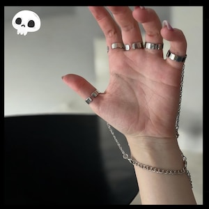 Może przedstawiać: Srebrna bransoletka na dłoń z wieloma pierścieniami połączonymi łańcuchem. Pierścienie są noszone na kilku palcach. Bransoletka ma wzór ogniw łańcucha. Tło jest w neutralnym kolorze.