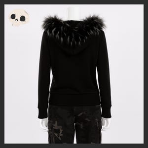 May include: A black hooded sweatshirt with a faux fur trim around the hood. The sweatshirt is displayed on a mannequin. The model is wearing camouflage cargo pants. A cartoon skull is in the upper left corner.