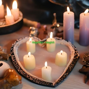 Pu&ograve; includere: Un vassoio a forma di cuore con cinque candele bianche accese al suo interno. Il vassoio &egrave; circondato da altre candele, tra cui candele viola, rosa e bianche. Il testo "Angelika's magic" &egrave; visibile sul vassoio.