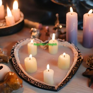 K&ouml;nnte beinhalten: Ein herzf&ouml;rmiges Tablett mit f&uuml;nf brennenden wei&szlig;en Kerzen. Das Tablett ist von anderen Kerzen umgeben, darunter violette, rosa und wei&szlig;e Kerzen. Der Text "Angelika's magic" ist auf dem Tablett sichtbar.