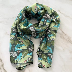 May include: A rectangular scarf featuring a repeating pattern of green frogs, blue lily pads, white flowers, dragonflies, and small bees against a dark charcoal background. The scarf has a thin black border along the edges and a smooth texture.