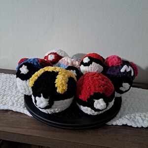 May include: A collection of handmade crochet balls in various colors, including red, black, white, blue, and yellow. The balls are arranged on a black plate, resting on a wooden surface. The balls have a round shape and are made of a soft, textured yarn.