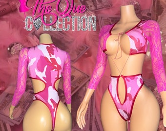 May include: Pink and white camouflage bodysuit with long pink lace sleeves. The bodysuit has a cut-out design on the back and front. The text "The Que Collection" is visible in the background.