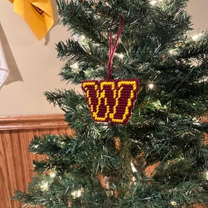 May include: A handmade Christmas tree ornament featuring a maroon and gold design. The ornament is shaped like a stylized "W" and is hanging from a red string. The ornament is displayed on a decorated Christmas tree with lights.