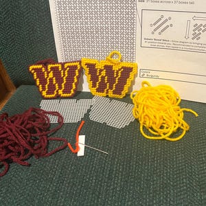 May include: Needlepoint craft kit featuring two burgundy and gold "W" designs. Includes yarn, plastic canvas, a needle, and instructions. The instructions show how to do the Gobelin 'Boxed' Stitch.