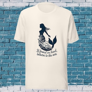 Puede incluir: Camiseta color crema con una silueta de sirena azul marino con cabello suelto y cola de pez. El diseño de la sirena incorpora elementos de coral y algas marinas. Debajo de la imagen está el texto: "If found on land, return to the sea."