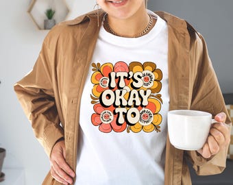Op de afbeelding: Wit T-shirt met de tekst "IT'S OKAY TO" in een retro lettertype, omringd door bloemmotieven in oranje, perzik en bruin. Het T-shirt wordt gedragen.