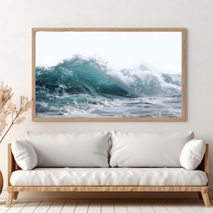 May include: A framed print of a crashing ocean wave in shades of teal and white. The artwork is displayed above a white sofa with wooden legs, creating a coastal theme. Decorative vase with dried plants.