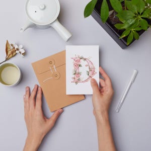 Puede incluir: Una tarjeta blanca con una letra D floral rosa en ella. La tarjeta est&aacute; siendo sostenida por una mano y est&aacute; encima de un sobre marr&oacute;n.
