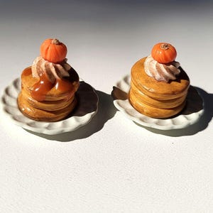 May include: Two miniature pancake stacks, each topped with whipped cream, a small orange pumpkin, and caramel sauce, sit on white scalloped-edge plates. The pancakes are golden brown, and the scene is lit by bright sunlight.