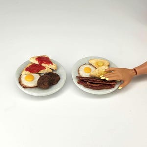 May include: Two plates of miniature breakfast food. One plate has toast with red jam, a fried egg, and a brown patty. The other plate has toast with butter, a fried egg, and strips of bacon. A hand is reaching for the second plate.