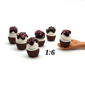 May include: Miniature chocolate cupcakes with white frosting and dark chocolate drizzle, topped with red sprinkles. The cupcakes are arranged on a white surface. The text "1:6" is visible.
