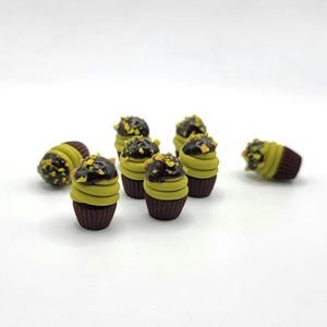 May include: A collection of miniature cupcakes with chocolate frosting and pistachio toppings. The cupcakes have a brown base, a light green frosting layer, and are arranged on a white surface. The image showcases a dessert theme.