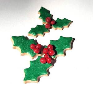 May include: Close-up of four holly leaf decorations. Each leaf is green with a glittery finish and a light brown border. Red berry clusters are attached to each leaf, creating a festive holiday theme.