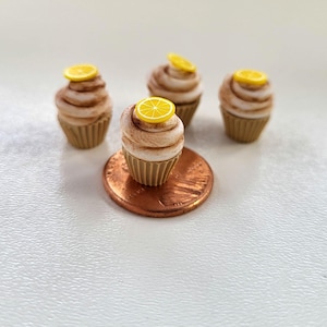 May include: Four miniature lemon cupcakes with yellow frosting and a lemon slice on top. The cupcakes are sitting on a white surface with a copper penny in front of one of the cupcakes.