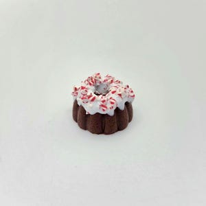 May include: A miniature dark brown bundt cake topped with white icing and small red and white peppermint candy pieces. The cake has a fluted, ring-shaped design with a hole in the center, featuring a textured surface and a festive appearance.