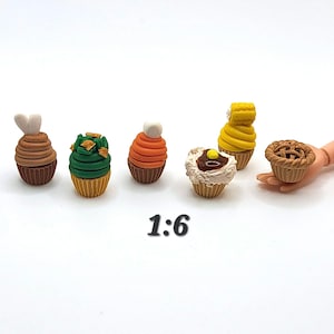 May include: A collection of six miniature cupcakes and a pie. The cupcakes feature various colors and toppings, including green frosting with sprinkles, orange frosting with a white ball, and yellow frosting. The pie is held by a hand.