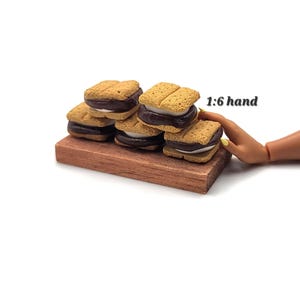 May include: A collection of four miniature s'mores, each featuring two tan graham cracker squares with a dark brown chocolate layer and a white marshmallow center, arranged on a rectangular wooden board with the text 1:6 hand visible.