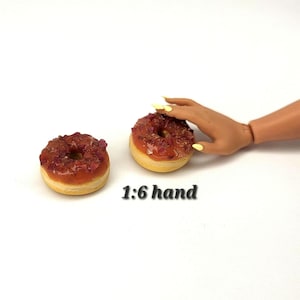 May include: Two miniature donuts with a light brown glaze and topped with reddish-brown sprinkles. The donuts are on a white surface. The text "1:6 hand" is below the donuts.