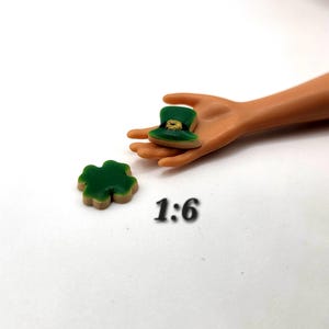 May include: Miniature green leprechaun hat with gold accents and a green shamrock. The hat is held in a hand, and a shamrock is on the surface. The text "1:6" is below the objects.
