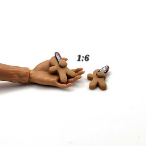 May include: Two brown gingerbread man-shaped charms with silver and red accents. One charm is held in a hand, and the other is placed separately. The text "1:6" is in the upper right corner.