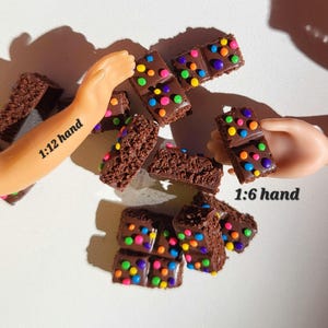 May include: Miniature chocolate brownies with colorful candy sprinkles. The brownies are arranged on a white surface, with a doll's hand holding some of them. The text "1:12 hand" and "1:6 hand" are visible.