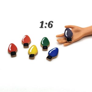 May include: Miniature colorful Christmas light bulbs in red, yellow, green, and blue, with a black base. One bulb is held by a hand. The number "1:6" is in the upper center. These are likely dollhouse or craft supplies.