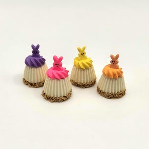 May include: Four miniature Easter-themed desserts. Each has a cream-colored base with a textured design, topped with colorful frosting and a bunny figure. Frosting colors include purple, pink, yellow, and orange. The bases sit on a brown, decorative edge.