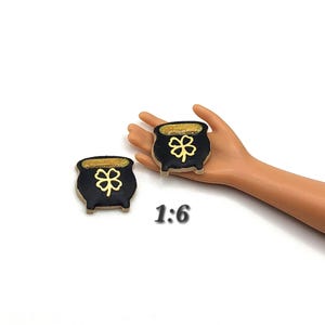 May include: Two black pot-shaped charms with gold shamrock designs and gold rims. The charms are on a white background. The text "1:6" is visible in the image.