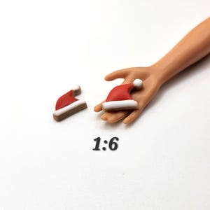 May include: Miniature red Santa hats with white trim and a white pom-pom. One hat rests on a hand, while another sits separately. The number "1:6" is printed in black below the hats.