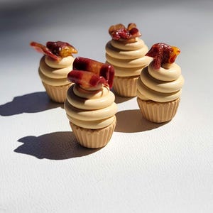 May include: Four miniature cupcakes with tan frosting and a topping that resembles bacon. The cupcakes sit on a white surface, with shadows cast by the sunlight. The cupcakes have a detailed, textured appearance.
