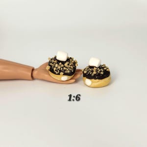 May include: Two miniature donuts with chocolate icing, chopped nuts, and a marshmallow topping. The donuts have a yellow base and are displayed on a white surface. The text "1:6" is visible.
