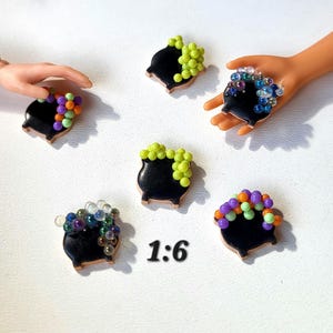 May include: Miniature black cauldron charms with brown trim, each topped with colorful beads. The charms are arranged on a white surface, with the text "1:6" visible. Some charms are held by a hand.