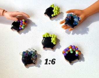 May include: Miniature black cauldron charms with brown trim, each topped with colorful beads. The charms are arranged on a white surface, with the text "1:6" visible. Some charms are held by a hand.