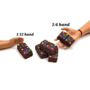 May include: Miniature chocolate brownies with colorful candy sprinkles. The brownies are arranged on a white surface, with two doll hands holding some of the brownies. The image is well-lit and the brownies appear to be freshly made.