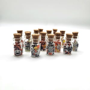 May include: A collection of small glass bottles with cork stoppers, filled with colorful candies. Each bottle features a Halloween-themed label with images of pumpkins, ghosts, and the text "Trick or Treat". The bottles are arranged on a white surface.