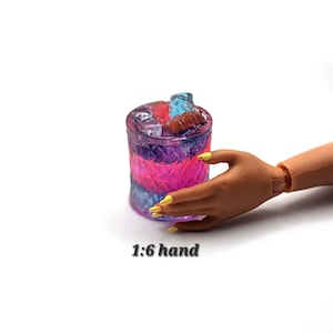 May include: A miniature tumbler glass featuring a textured diamond pattern on the exterior, filled with a vibrant pink and blue liquid and translucent ice cubes. The glass is positioned next to a 1:6 scale hand with yellow painted fingernails.