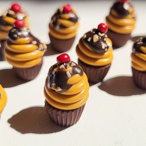 May include: A collection of miniature cupcakes with yellow frosting, chocolate drizzle, and a red cherry on top. The cupcakes sit in brown paper cups. The image is well-lit, highlighting the details of the desserts.