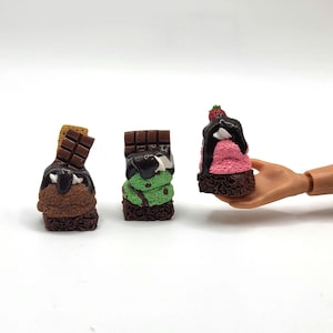 May include: Miniature desserts: chocolate, mint, and strawberry ice cream sundaes with chocolate sauce and chocolate bar toppings. One sundae is held by a doll hand. The sundaes are on a white background.
