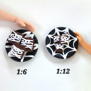 May include: Two black plates with white spiderweb designs, each holding miniature mummy brownies. The brownies are decorated with white icing bandages and edible eyes. The text "1:6" and "1:12" are below the plates, indicating scale.