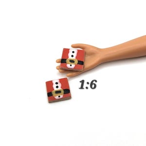 May include: Two miniature Santa Claus suit squares. Each square is red with a white collar, a black belt with a gold buckle, and two black buttons. The text "1:6" is next to the squares.