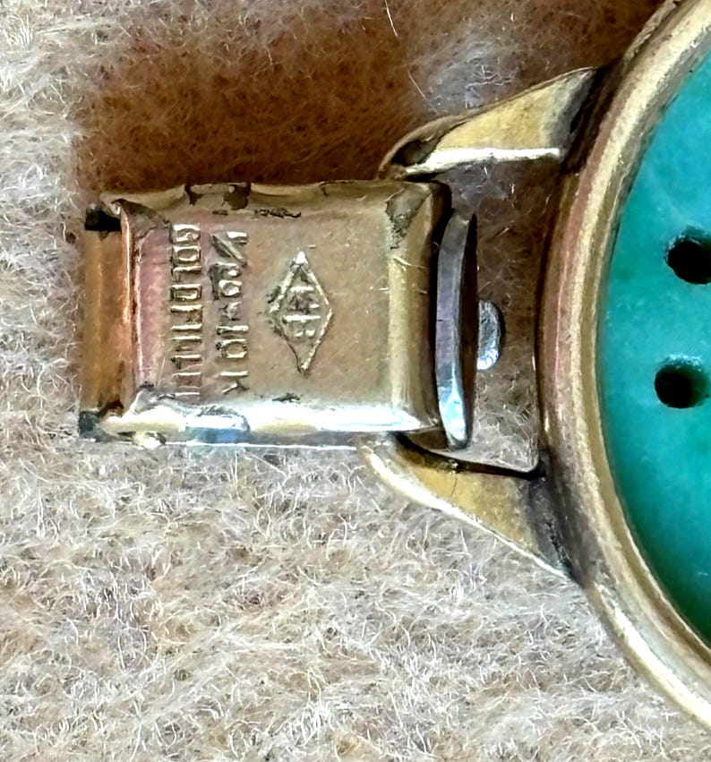 May include: Close-up of a gold-toned bracelet clasp with the text "1/20 10K Gold Filled" and a diamond-shaped hallmark. The bracelet features a turquoise-colored stone.