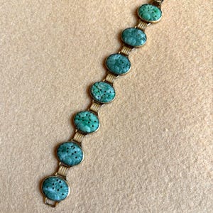 May include: A vintage bracelet featuring oval, carved jade stones in a vibrant green hue. The jade stones are set in a gold-toned metal, connected by delicate links. The bracelet is displayed on a neutral-toned surface.