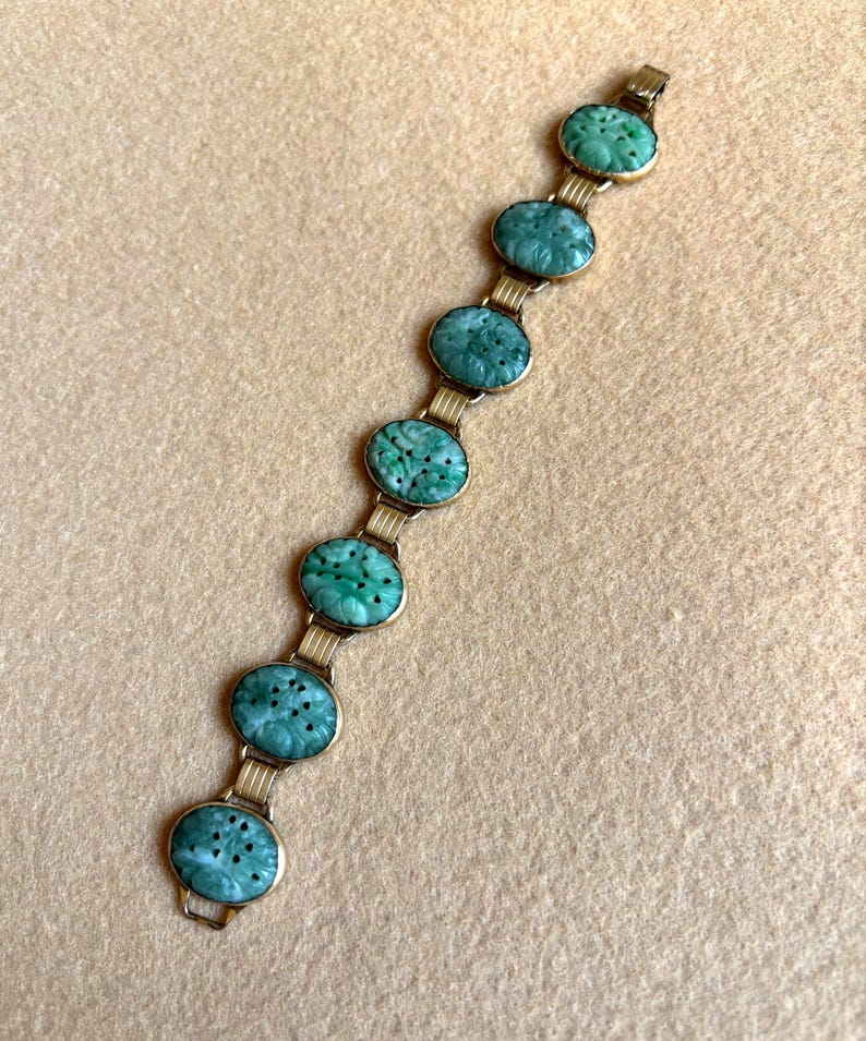 May include: A vintage bracelet featuring oval, carved jade stones in a vibrant green hue. The jade stones are set in a gold-toned metal, connected by delicate links. The bracelet is displayed on a neutral-toned surface.