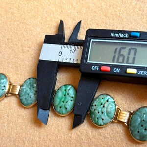 May include: A gold bracelet with oval, carved jade stones. The bracelet is being measured with a digital caliper, showing a measurement of 16.0 mm. The jade stones are a light green color.