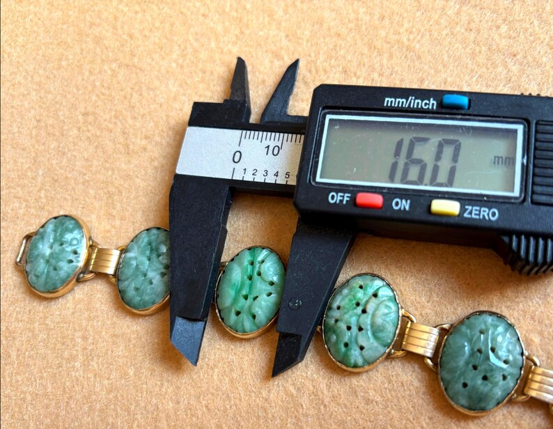 May include: A gold bracelet with oval, carved jade stones. The bracelet is being measured with a digital caliper, showing a measurement of 16.0 mm. The jade stones are a light green color.