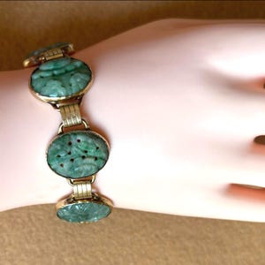 May include: A gold-toned bracelet with oval, carved jadeite links. The jadeite is a rich green color, and the carvings appear to be floral or nature-inspired. The bracelet is displayed on a light-toned skin.