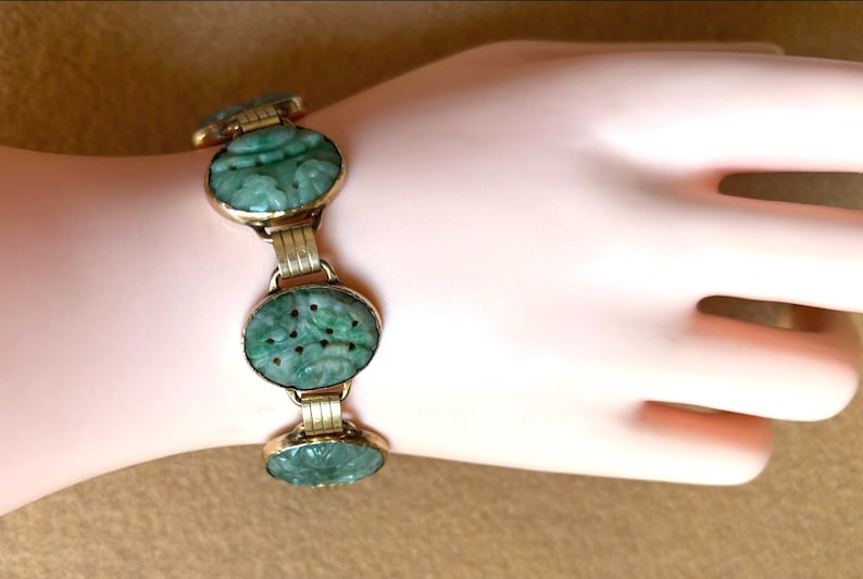 May include: A gold-toned bracelet with oval, carved jadeite links. The jadeite is a rich green color, and the carvings appear to be floral or nature-inspired. The bracelet is displayed on a light-toned skin.