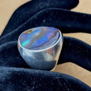 May include: A silver ring with a large, oval abalone shell stone. The iridescent shell displays shades of blue, green, and purple. The ring is set on a dark blue velvet surface.