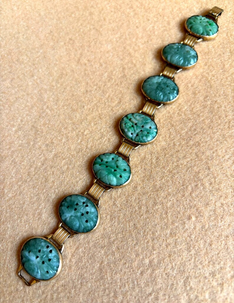 May include: A vintage bracelet featuring oval, carved jade stones in a vibrant green hue. The jade stones are set in a gold-toned metal frame and connected by gold links. The bracelet is displayed on a neutral-toned surface.